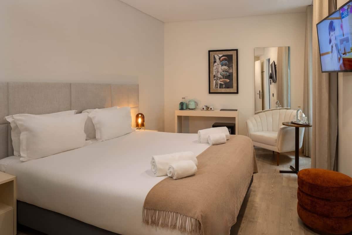 A hotel room with a large bed, a bedside table with a lamp, a desk with a chair, a small round ottoman, and a TV mounted on the wall.