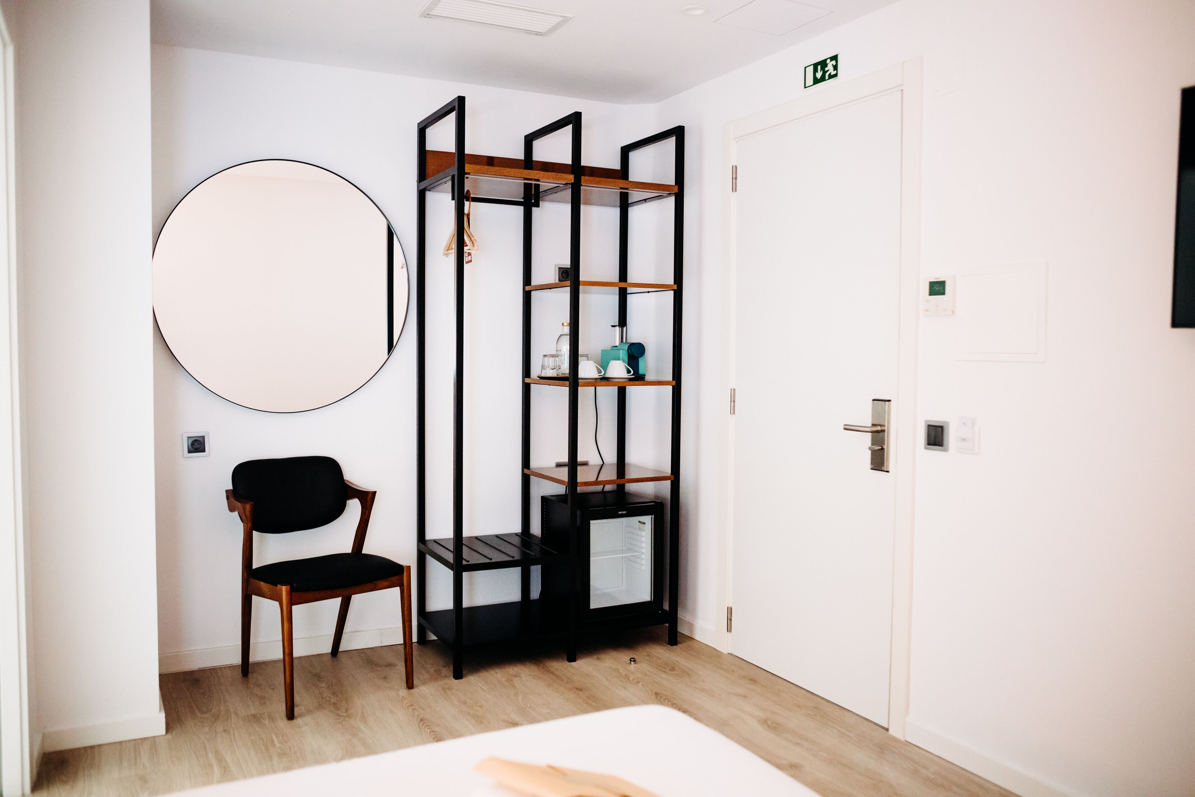 A room with a bed, a chair, a round mirror, a black metal shelving unit, and a white door with an exit sign.
