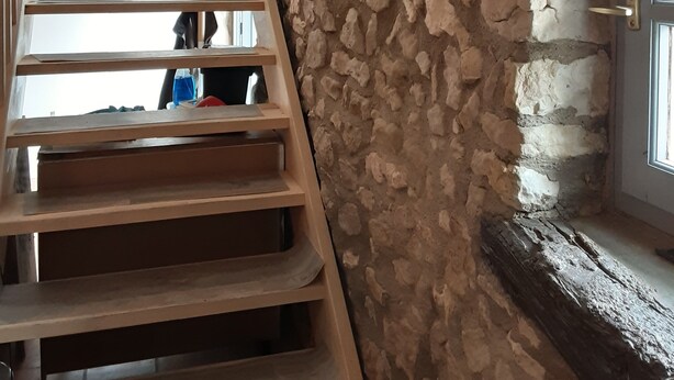 Una escalera de madera con peldaños que suben a una plataforma, un muro de piedra y una puerta con un ventanal.