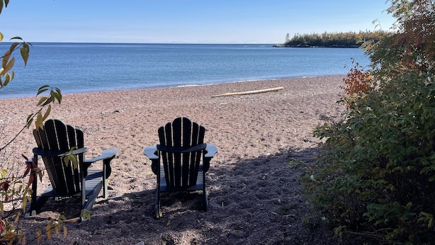 Zwei Adirondack-Stühle, die auf einen Strand mit Blick auf Wasser und Bäume in der Ferne zeigen.