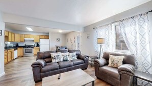 A modern living room with a sofa, armchairs, and a coffee table. A kitchen with wooden cabinets and appliances is visible in the background. - Great Two King Bedroom Spot Close to the Airport (Brighton)