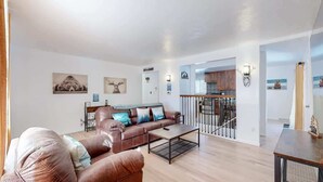A spacious living room with a brown leather sofa, a coffee table, and artwork on the walls. A staircase leads to a kitchen area. - Spacious Arvada Home with King Beds and Office (Arvada)