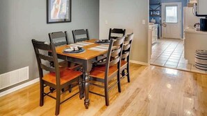 Een eethoek met een houten tafel en stoelen, een ingelijst beeld aan de muur en een keuken op de achtergrond.