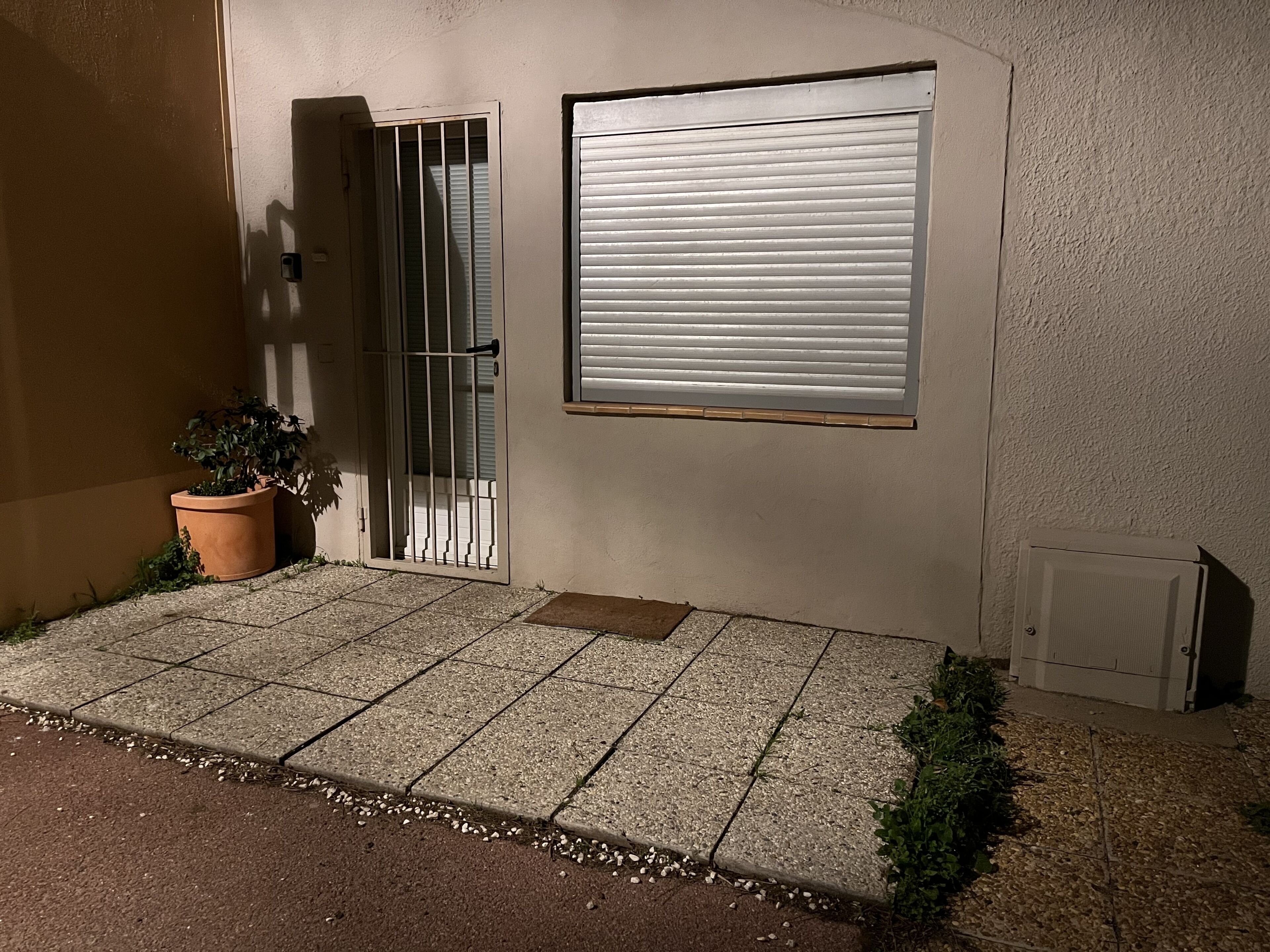 A building facade with a closed metal gate, a potted plant, and a window with horizontal blinds.