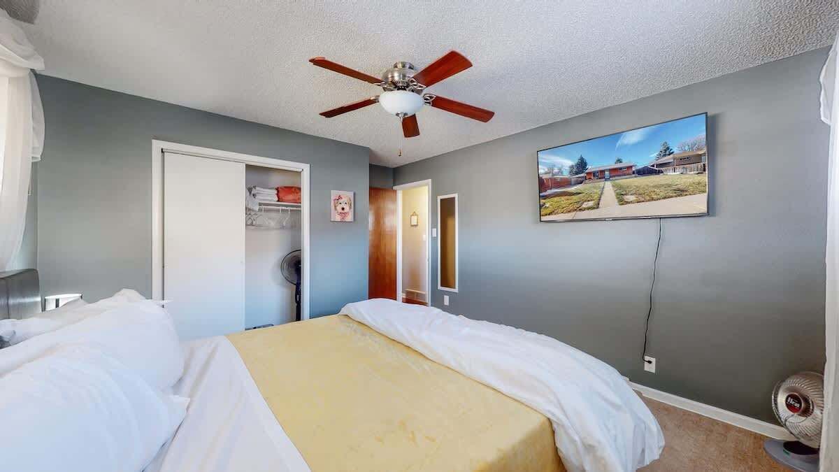 Un dormitorio con cama, ventilador de techo, armario, puerta y televisor montado en la pared.