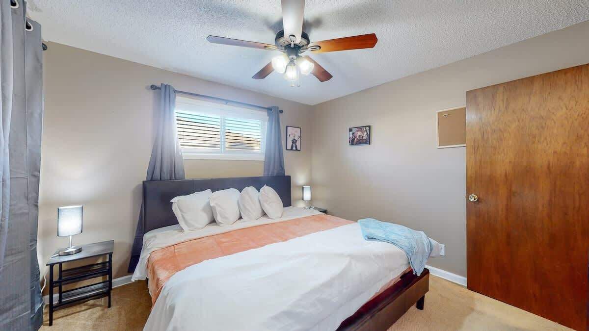 Un dormitorio con cama, lámparas de noche, ventilador de techo, ventana con persianas y puerta de madera.