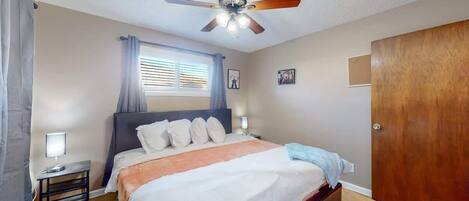 Un dormitorio con cama, lámparas de noche, ventilador de techo, ventana con persianas y puerta de madera.