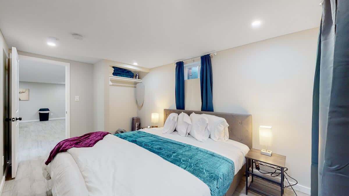 A bedroom with a bed, bedside table, and a nightstand. There is a window with curtains, a door, and a small shelf with items.