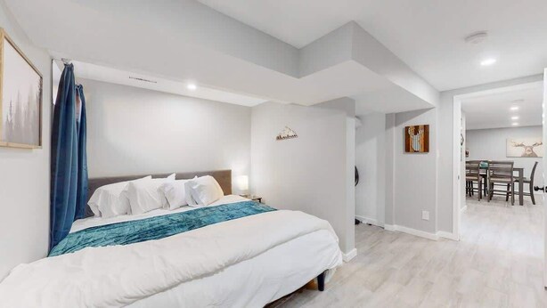 Une chambre moderne avec un grand lit, une table de chevet et une commode. On aperçoit un coin repas à l’arrière-plan.