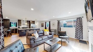 A modern living room with a sofa, coffee table, and armchairs. A kitchen with dark cabinets and a dining area with a table and chairs is visible in the background. - Spacious Family Retreat in Denver (Denver)