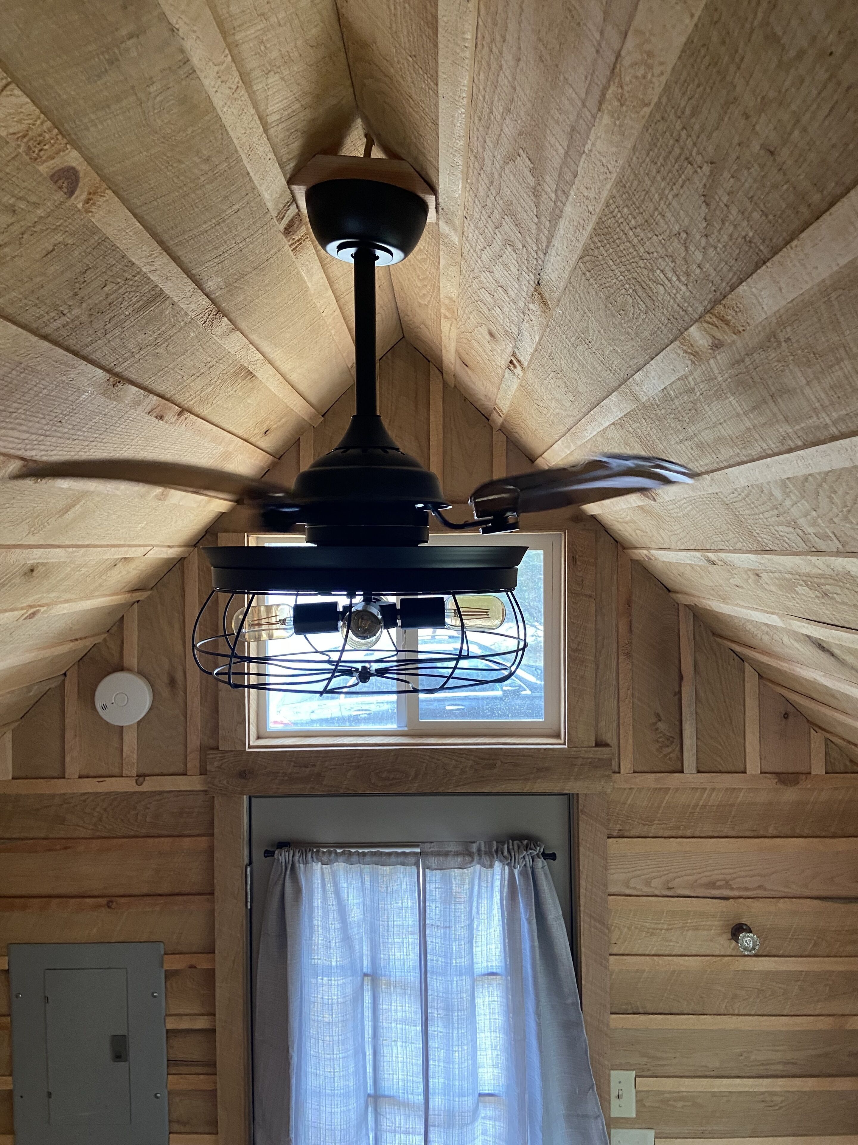 Ein Zimmer mit schrägen Wänden, einem Deckenventilator, einem Fenster mit Vorhängen und einer Tür mit Lichtschalter.