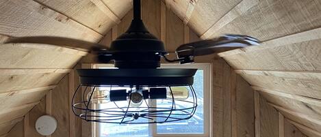 Ein Zimmer mit schrägen Wänden, einem Deckenventilator, einem Fenster mit Vorhängen und einer Tür mit Lichtschalter.