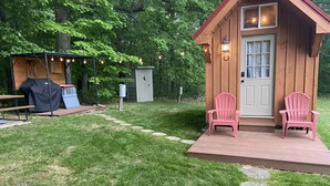 A wooden cabin with two pink Adirondack chairs on a deck, a covered grill, and a stone pathway leading to the door.