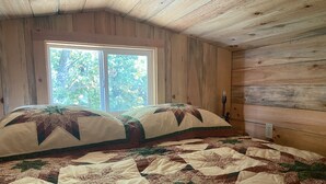 A bedroom with a large bed, a window with a view of trees, wooden walls, and a nightstand with a lamp.
