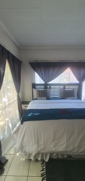A bedroom with a bed, curtains, a window, and a nightstand. - Anopam Guest House  (Pretoria)