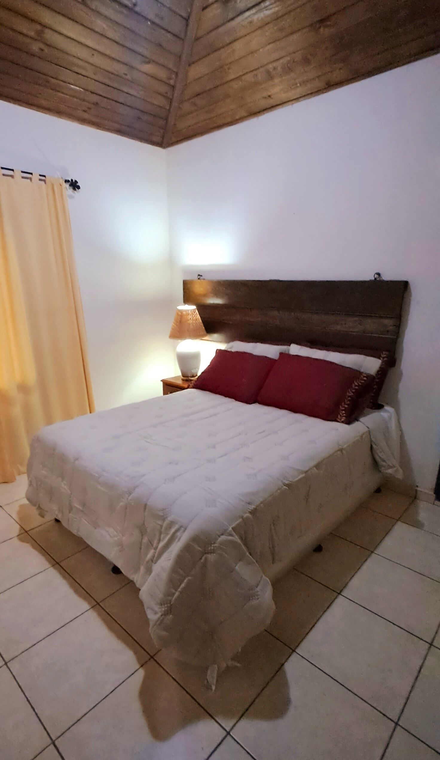 Una cama con cabecera de madera, una lámpara de noche y una cortina.