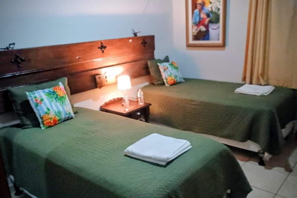 Quarto de hotel com duas camas de solteiro, uma mesa de cabeceira com abajur e um armário de madeira.