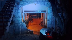 Entrada de um edifício com arco de pedra, uma escadaria à esquerda e interior iluminado em vermelho visível pelo arco.