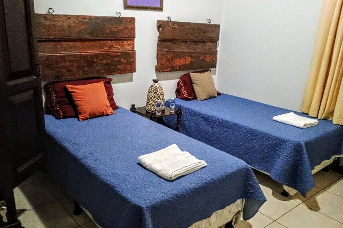 Quarto com duas camas, cada uma com colcha azul e toalha branca. Apresenta cabeceiras de madeira e uma pequena mesa com um vaso e uma garrafa.