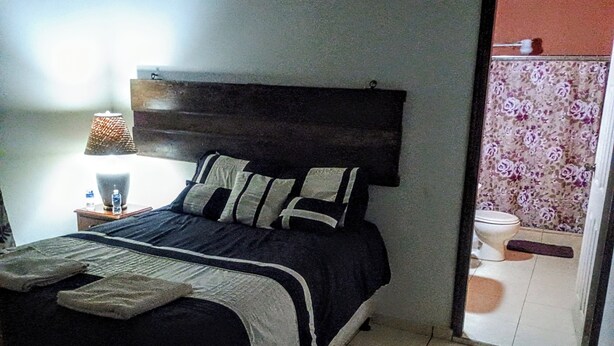 Quarto com cama, mesa de cabeceira com luminária e banheiro visível através de uma porta aberta.