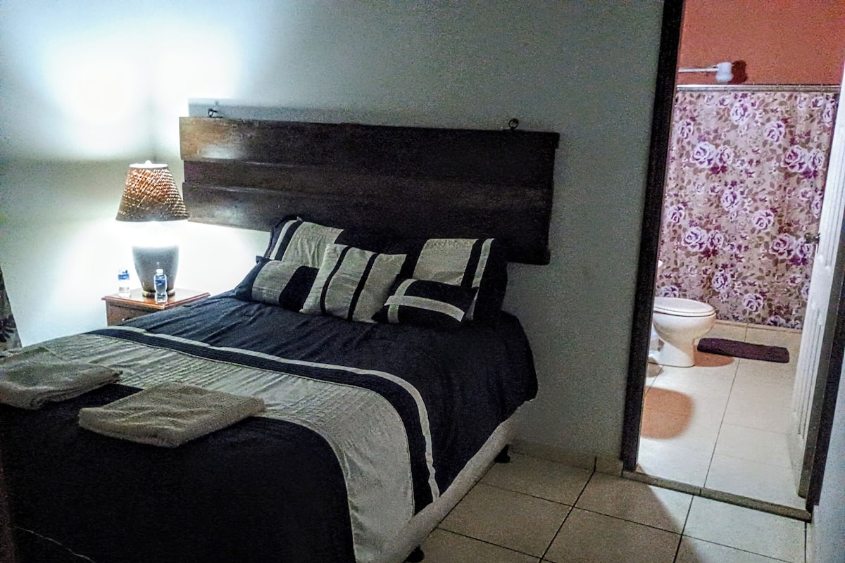 Quarto com cama, mesa de cabeceira com luminária e banheiro visível através de uma porta aberta.