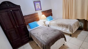 A room with two beds, a wooden wardrobe, a nightstand with a lamp, and a framed picture on the wall. - Villa Chelsey (Valle de Angeles)