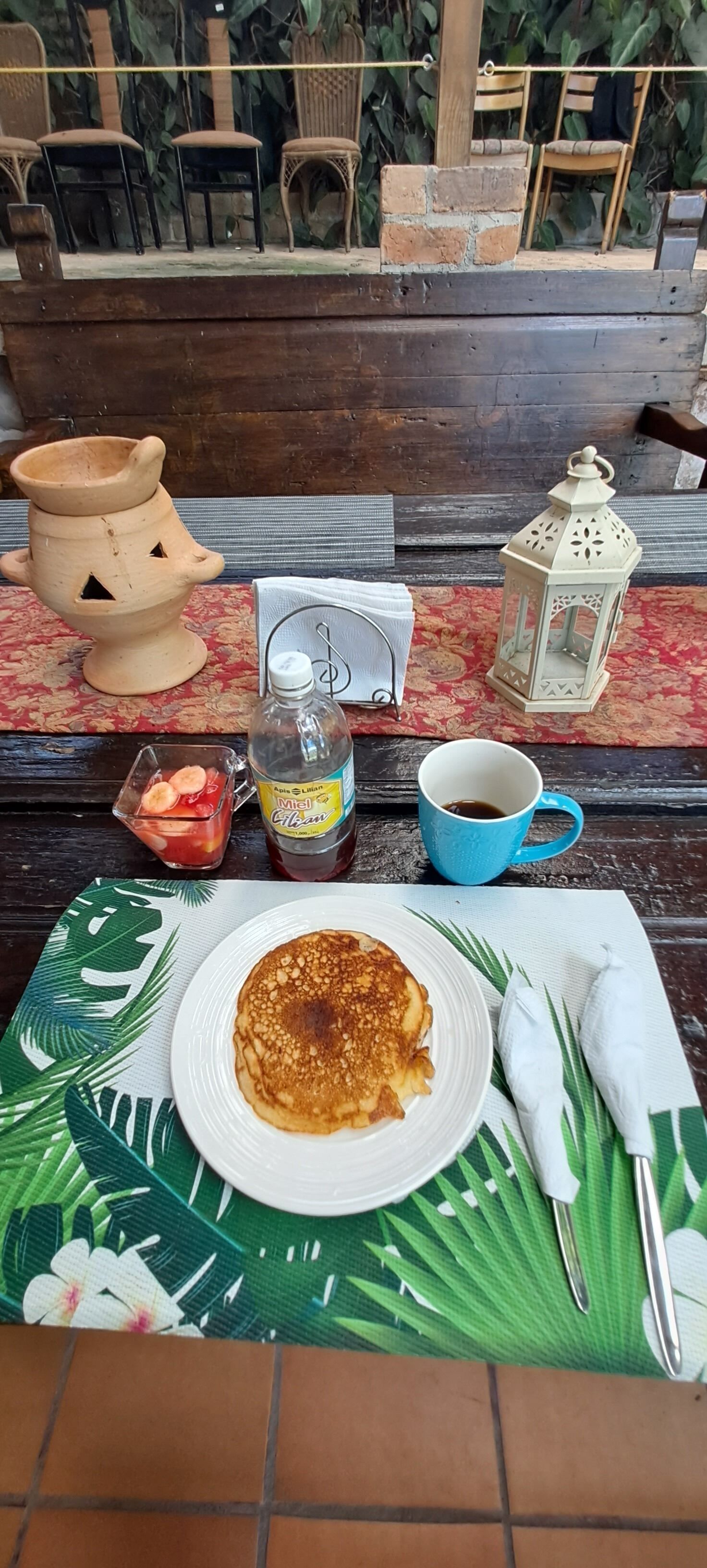 A table with a plate of pancakes, a bottle of syrup, a cup of coffee, a small bowl of fruit, a candle, and a lantern.