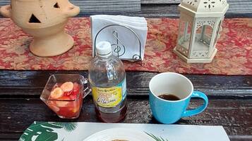 Una mesa con una pila de tortitas, una botella de almíbar, una taza de café, un bol pequeño de fruta, una vela y un farol.