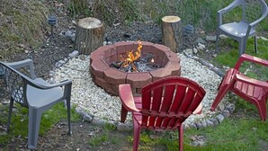 A fire pit surrounded by chairs and logs, set in a garden with gravel and greenery.