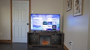 A living room with a flat-screen TV on a wooden stand, a fireplace below, and a door to the left.