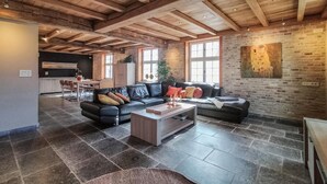 Un vaste espace de vie doté d’un plafond haut et de poutres apparentes en bois, d’un canapé en cuir noir, d’une table basse et d’un mur en briques orné d’un tableau.