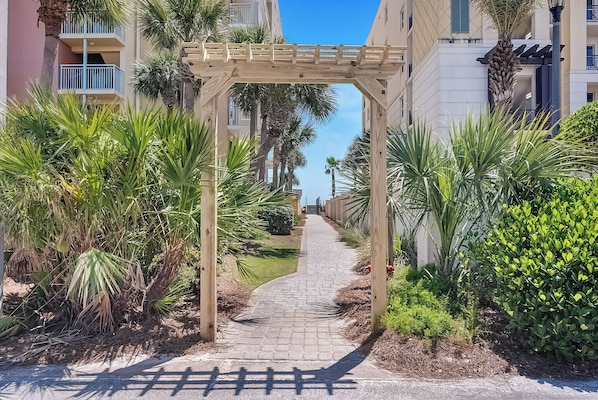 Un chemin menant à une pergola en bois dans un quartier résidentiel, avec des palmiers et un ciel dégagé.