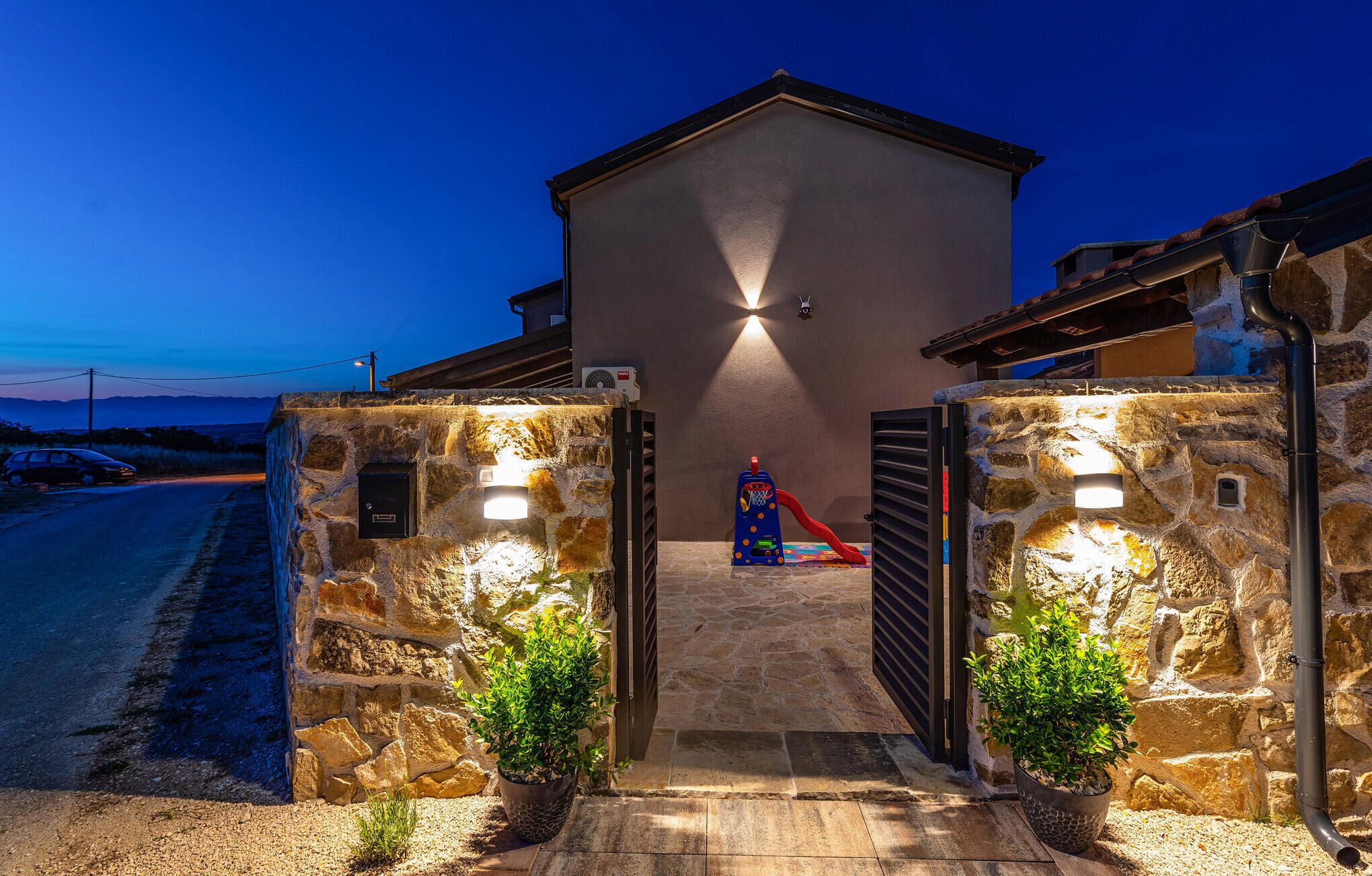 A stone entrance with a metal gate, a small play area with a slide, and a building with a sloped roof.