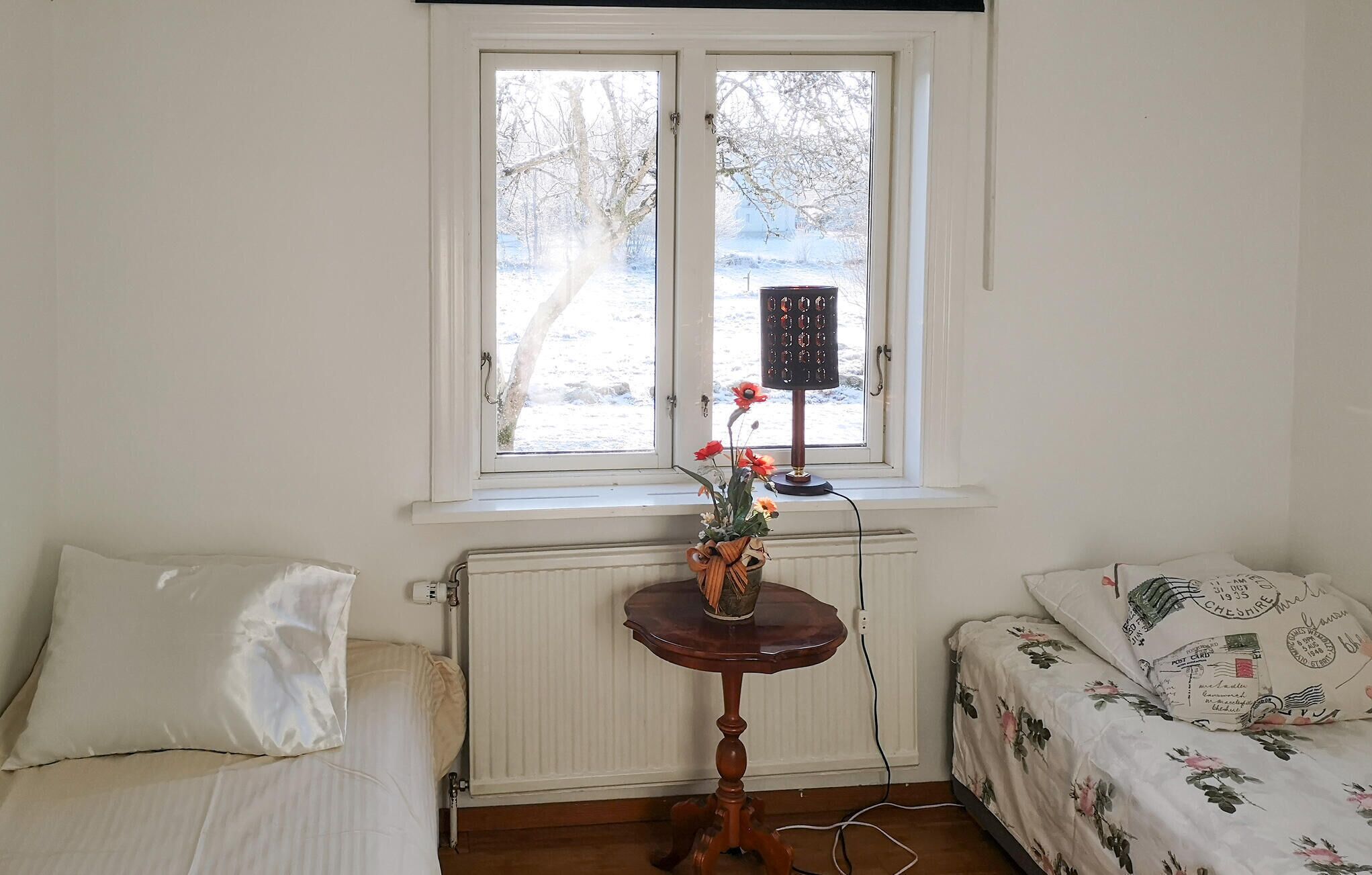 Une pièce comprenant un lit, une petite table avec un vase de fleurs, un radiateur et une fenêtre donnant sur des arbres, ainsi qu’une lampe.