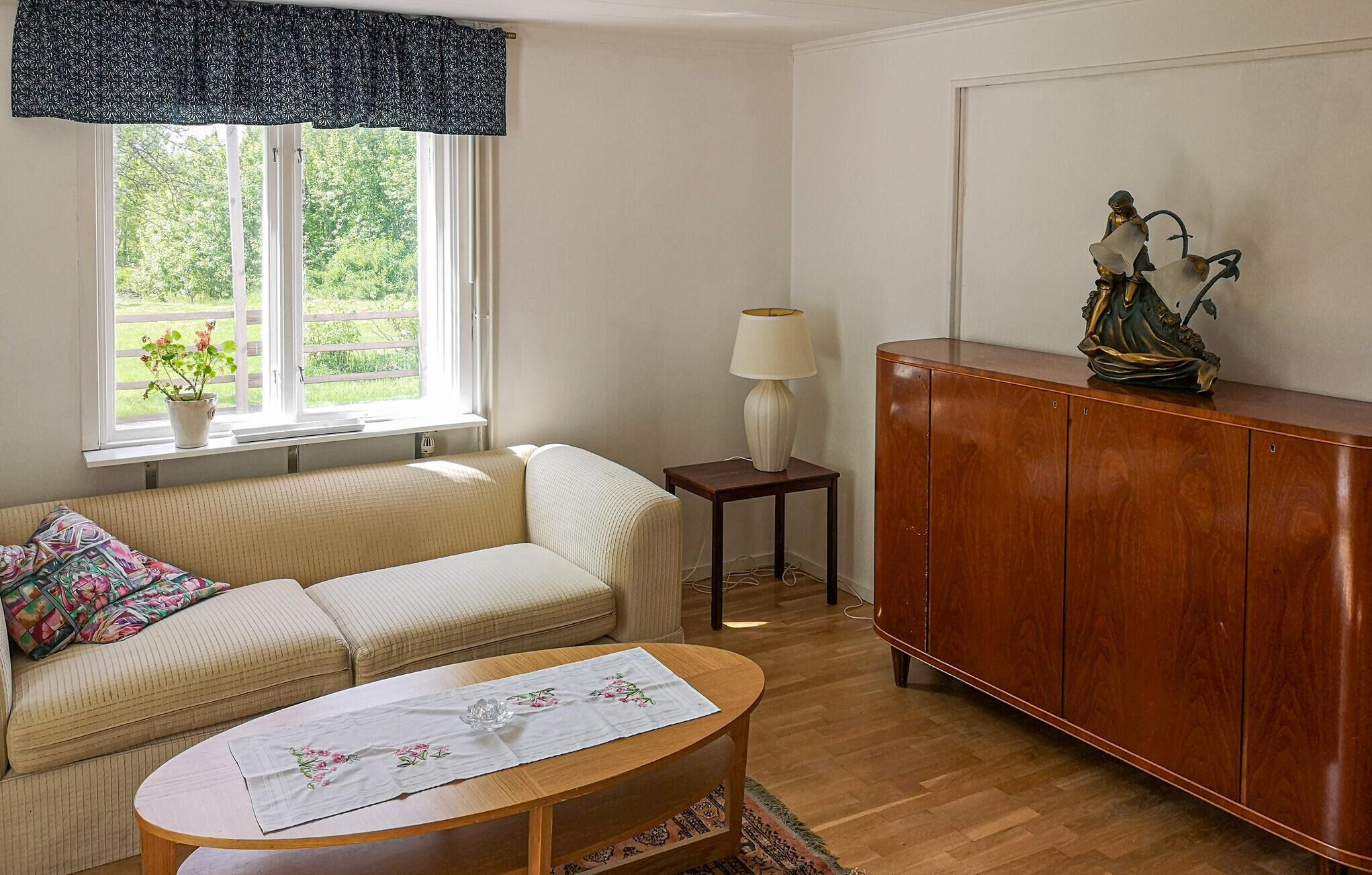 Un salon chaleureux avec un canapé, une petite table, un meuble, une lampe et une fenêtre donnant sur un paysage verdoyant.