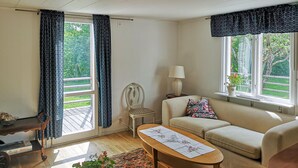 A cozy living room with a sofa, a wooden coffee table, a chair, and a lamp. There are two windows with curtains, a view of greenery outside, and a small table with a plant.