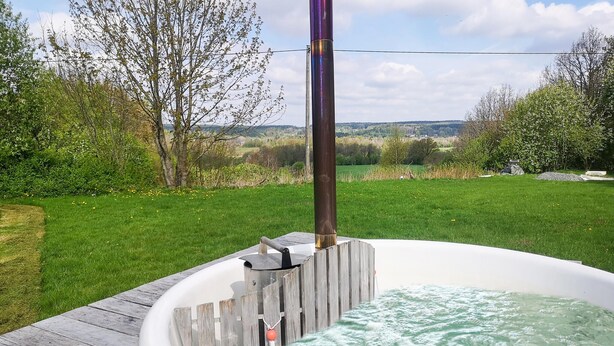 A hot tub with a wooden deck, surrounded by greenery and a clear view of the countryside.