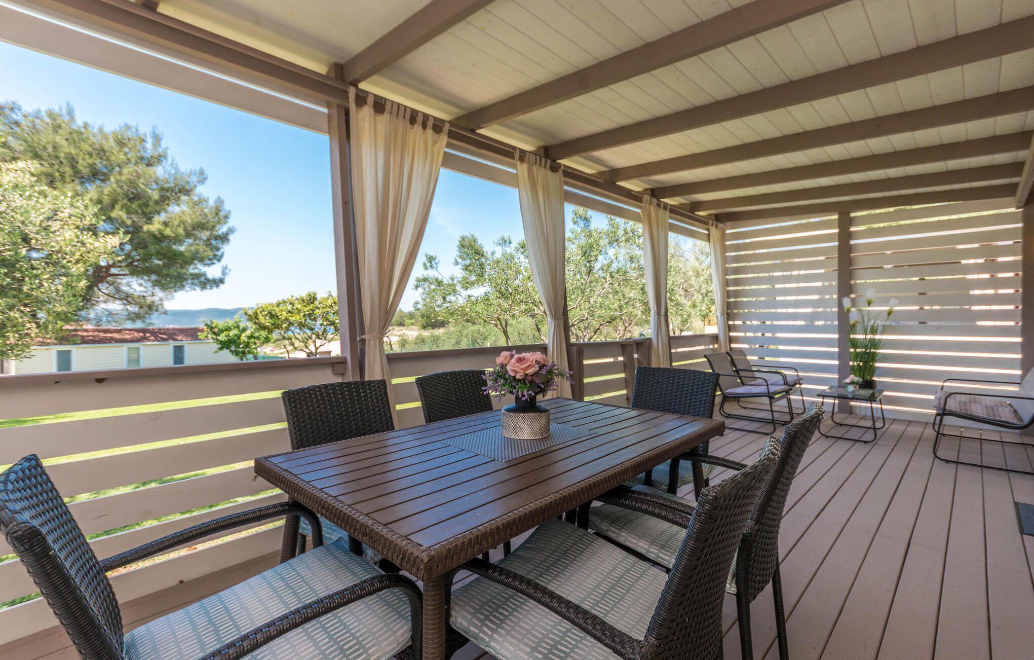 Une terrasse extérieure couverte, avec une table en bois et des chaises en osier, donnant sur une piscine et des arbres.