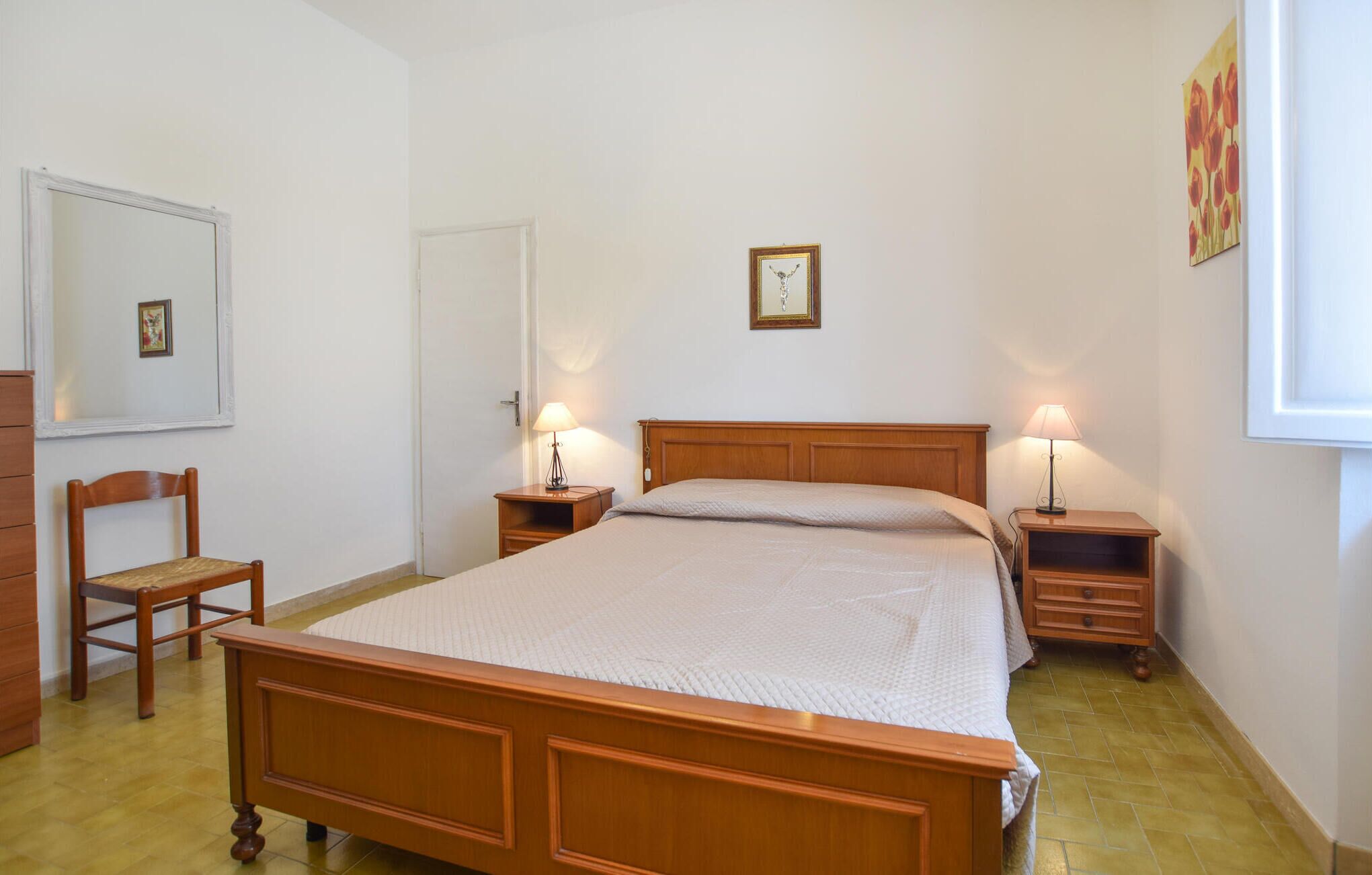 Une chambre à coucher avec un lit en bois, deux tables de chevet avec des lampes, une chaise, une commode et un tableau encadré au mur.