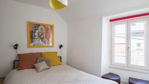 A bedroom with a bed, a painting, a window, and a view of buildings.