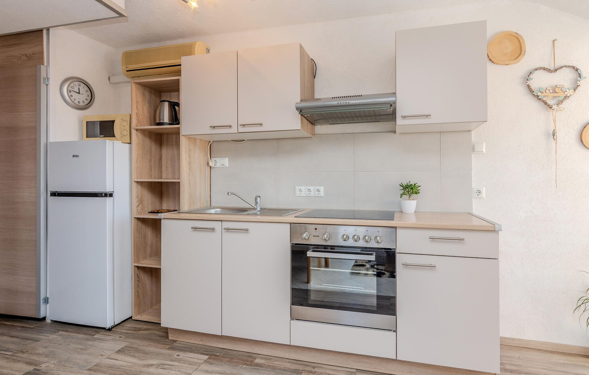 A compact kitchen with a built-in oven, a sink, and a stove. There is a clock on the wall and a small plant on the counter.
