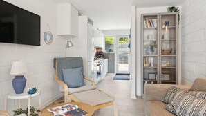 Un salon moderne comprenant un canapé, un fauteuil, une table basse avec des magazines, un téléviseur, une bibliothèque et une cuisine visible à travers une porte ouverte.