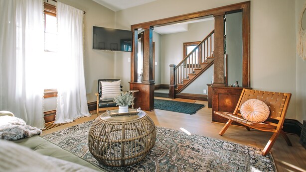 A spacious living room with a wooden floor, a large woven coffee table, a comfortable sofa, and a wooden chair. A staircase leads to an upper level, and a flat-screen TV is mounted on the wall. - 3 Min to downtown & Falls Park, 10 min to Airport - Clean King Bed Home (Sioux Falls)