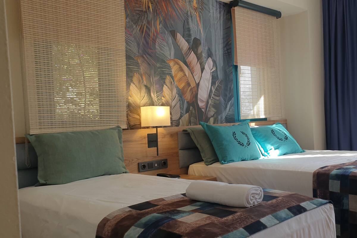 Een hotelkamer met twee bedden, een groot raam met zonwering en uitzicht op de omgeving.