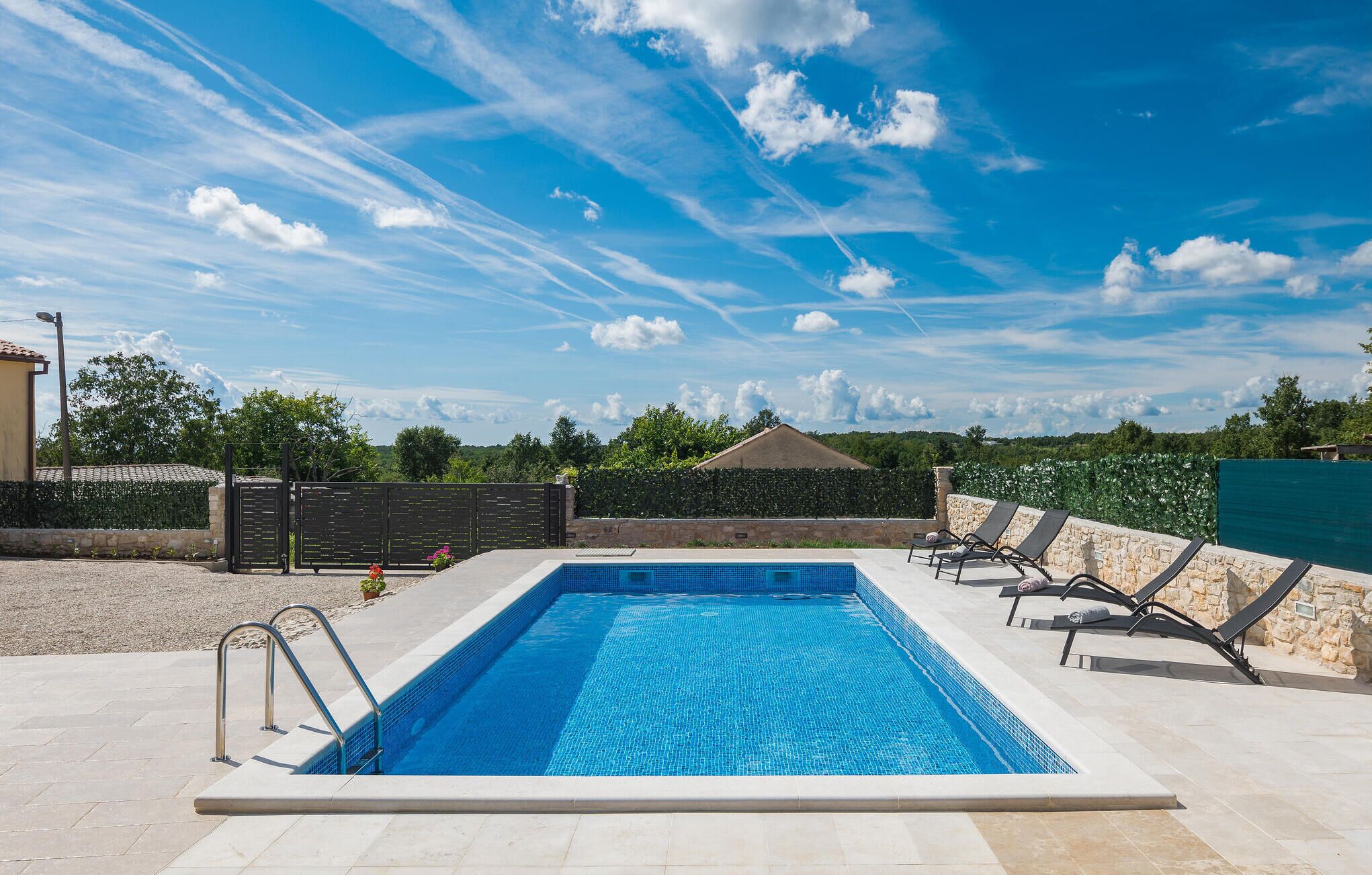 A rectangular swimming pool with a tiled edge, surrounded by a stone wall and a hedge. There are lounge chairs and a metal ladder leading into the pool.