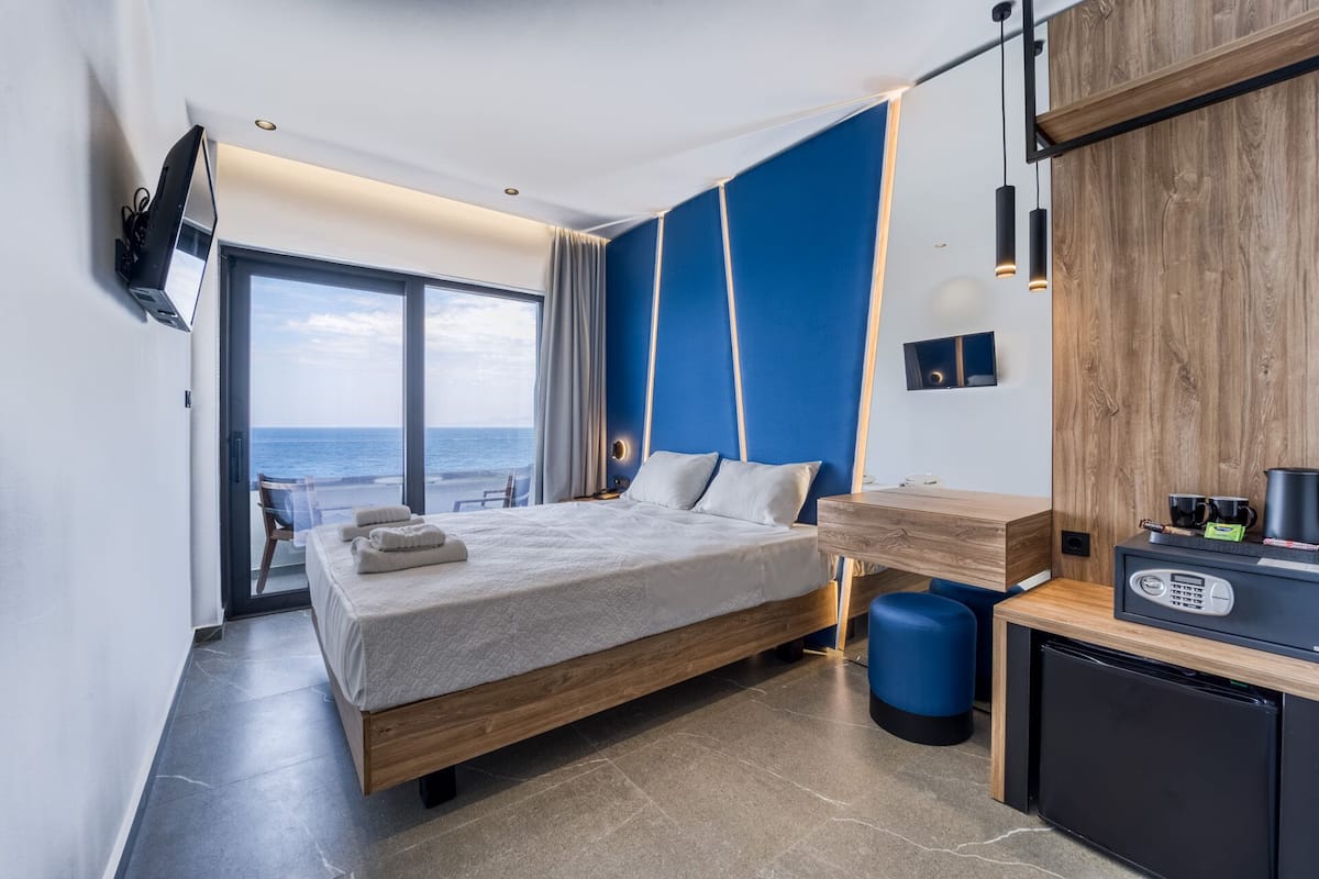 A modern hotel room with a large bed, a desk with a refrigerator, and a view of the sea.