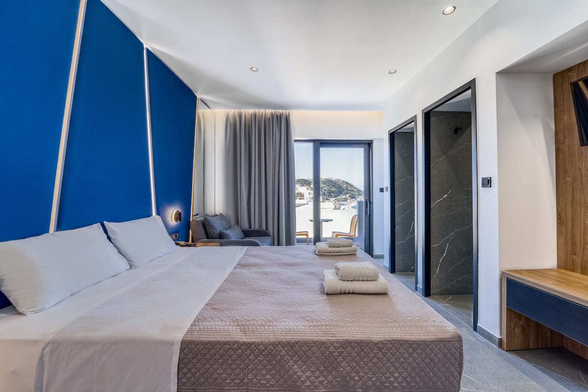 A modern hotel room with a large bed, a seating area with a view, and a sliding glass door leading to a balcony.
