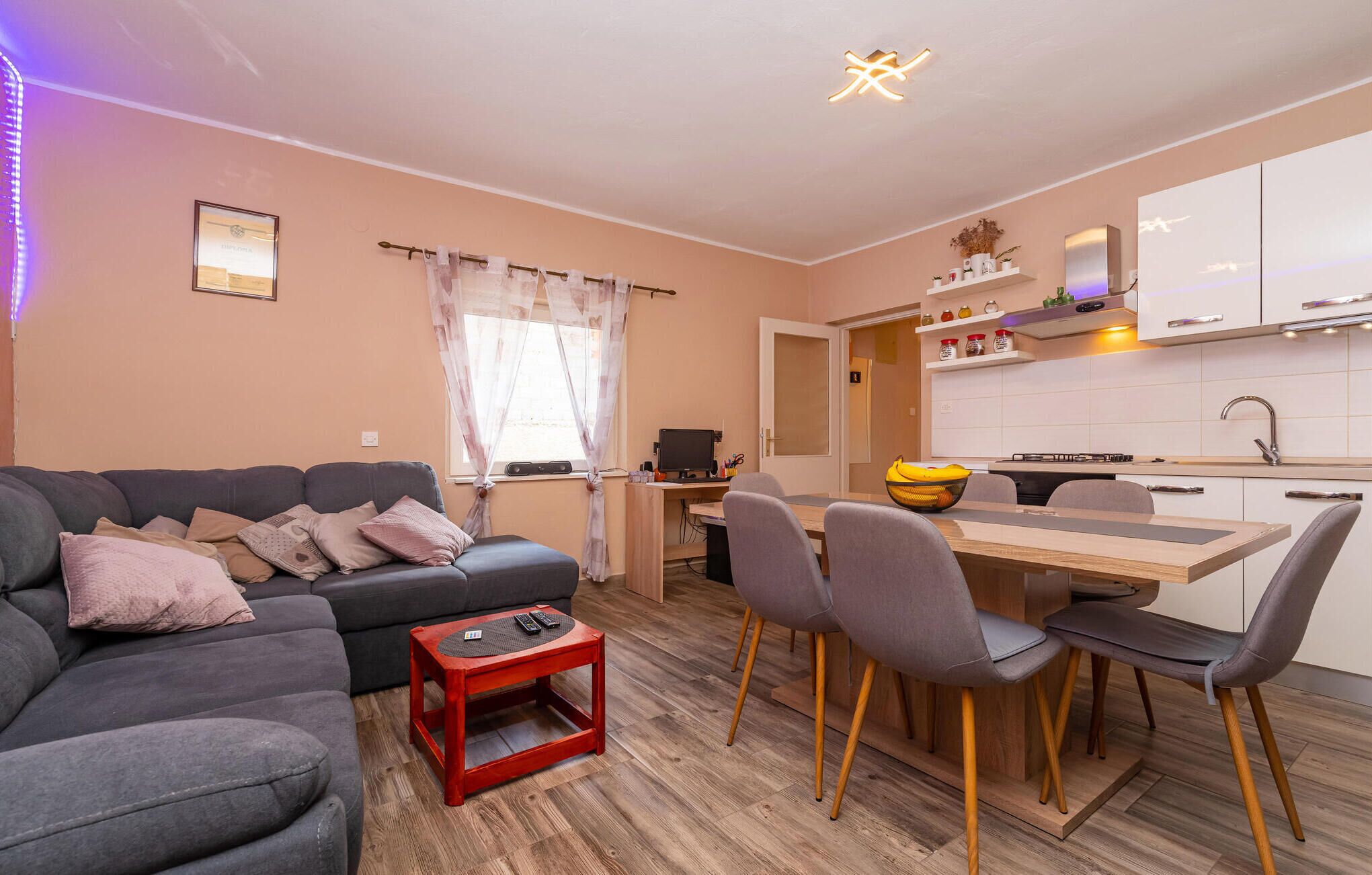 Uma sala de estar moderna com um sofá cinzento, uma mesa de centro vermelha e um espaço de jantar com uma mesa de madeira e cadeiras cinzas.