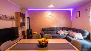 A cozy living room with a stone fireplace, a large dark sofa, a dining table with a fruit bowl, and a TV. - Gorgeous home in Pridraga with WiFi (Pridraga)