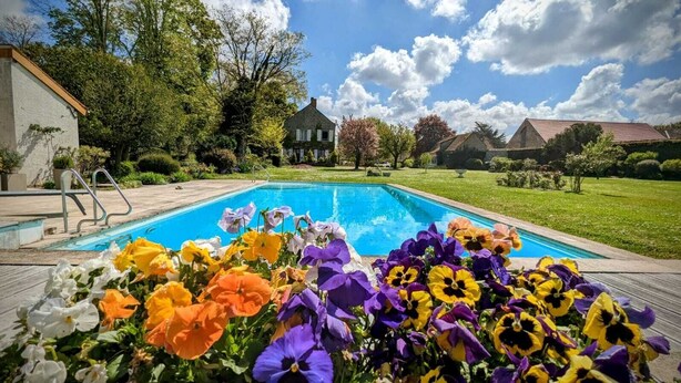 Une piscine entourée de fleurs aux couleurs vives, avec une maison et une végétation luxuriante en arrière-plan.